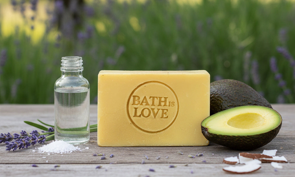 Bar of soap with 'Bath is Love' branding, a bottle of liquid, and an avocado on a wooden surface with a blurred natural background.