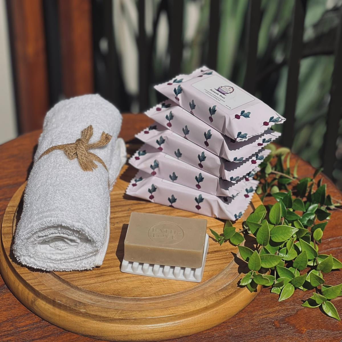 Stack of folded towels, a bar of soap, and a scrubber on a wooden surface with a blurred natural background.
