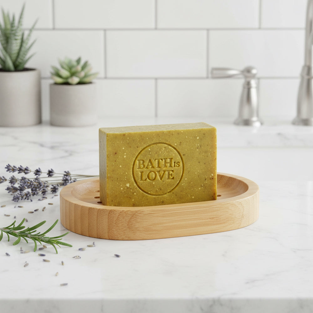 Bar of soap with 'Bath is Love' branding on a wooden soap dish, surrounded by lavender and rosemary on a marble countertop.