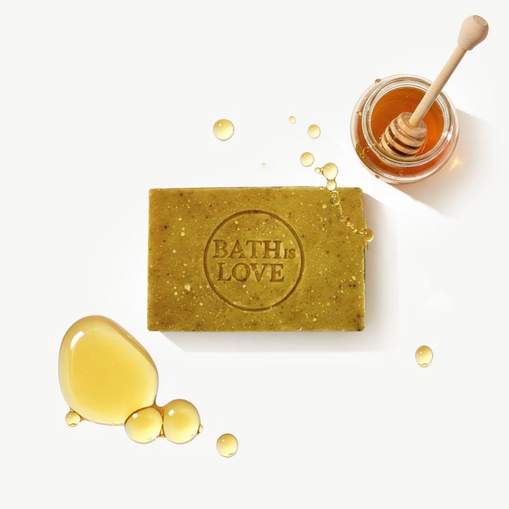 Bar of soap with 'Bath is Love' branding next to honey and a honey dipper on a white background