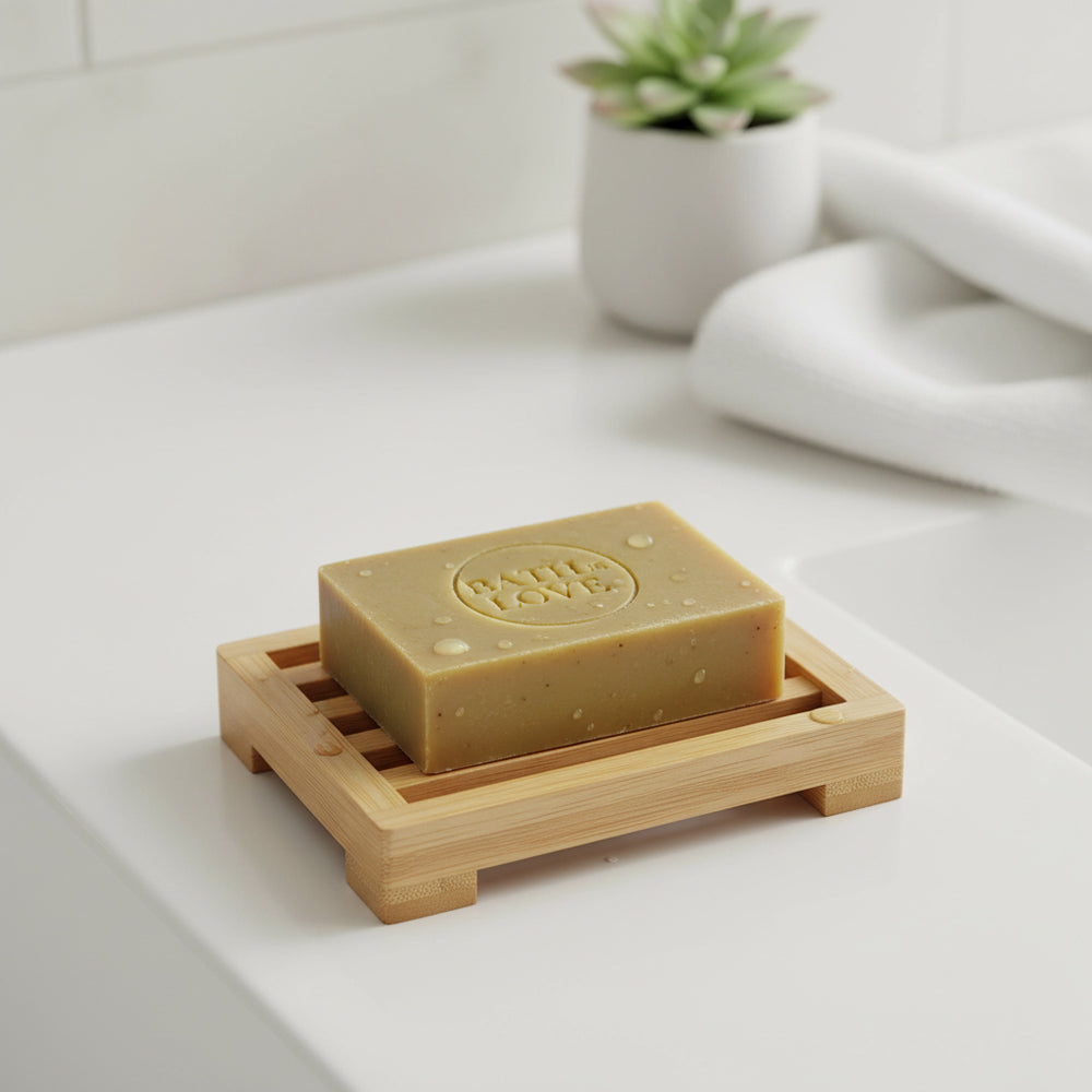 Bar of soap on a wooden soap dish with a plant and towel in the background