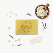 Bar of soap with 'Bath is Love' text, surrounded by lavender flowers and a bowl of cream on a white background.