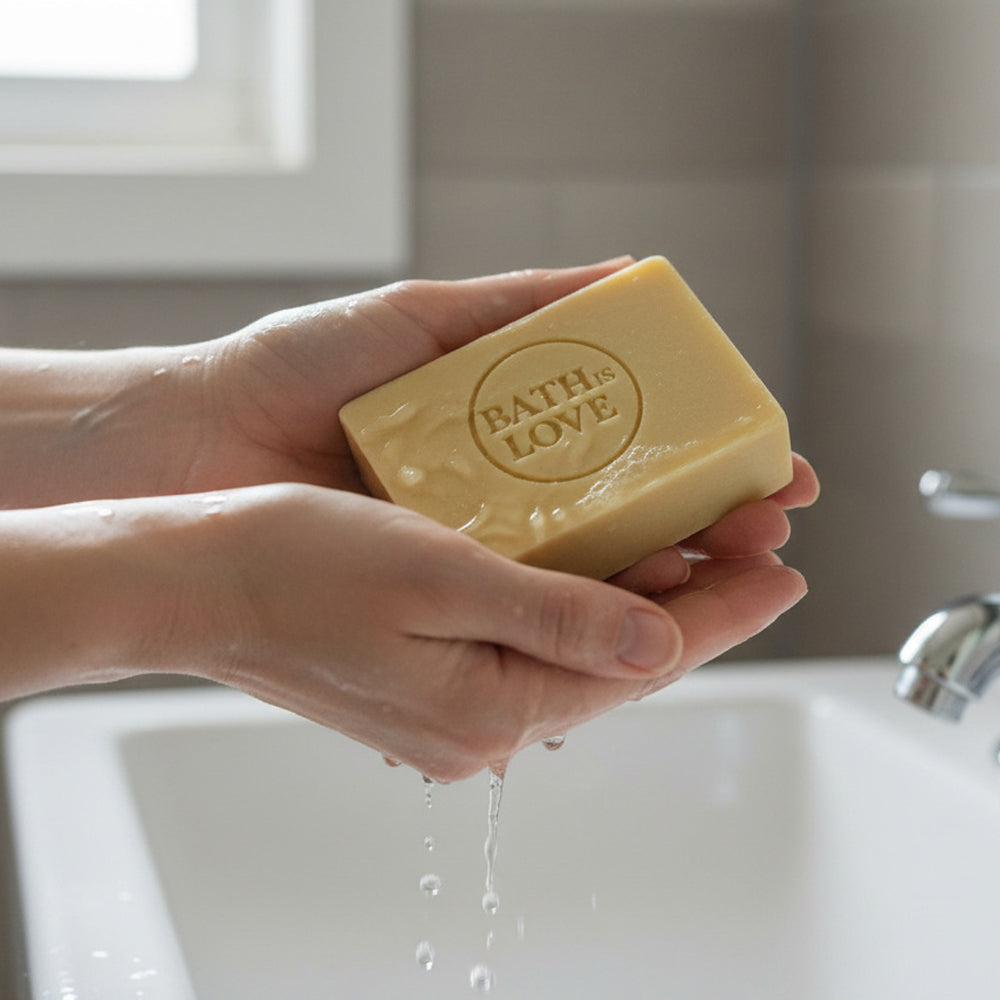 Hand holding a bar of soap labeled 'Bath is Love' over a sink.