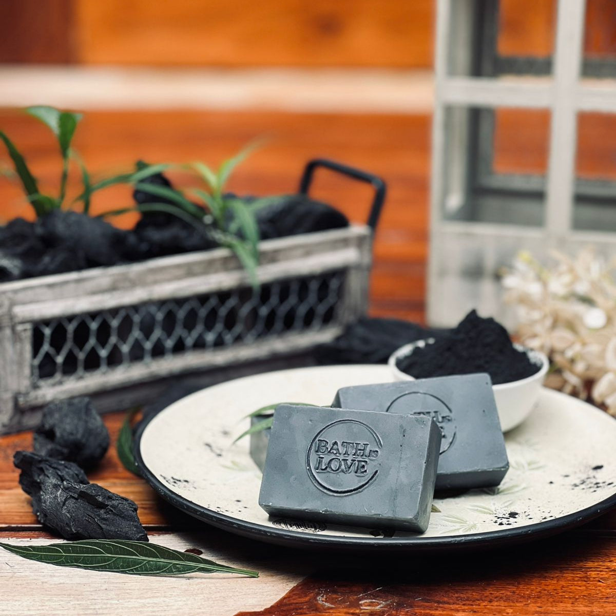 Black soap bars with 'Bath & Love' branding on a plate, surrounded by charcoal and plants.